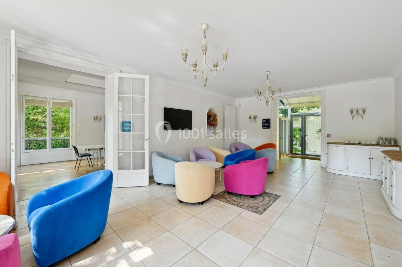 Salon lumineux avec fauteuils colorés, carrelage clair, lustres et vue sur un espace extérieur verdoyant.