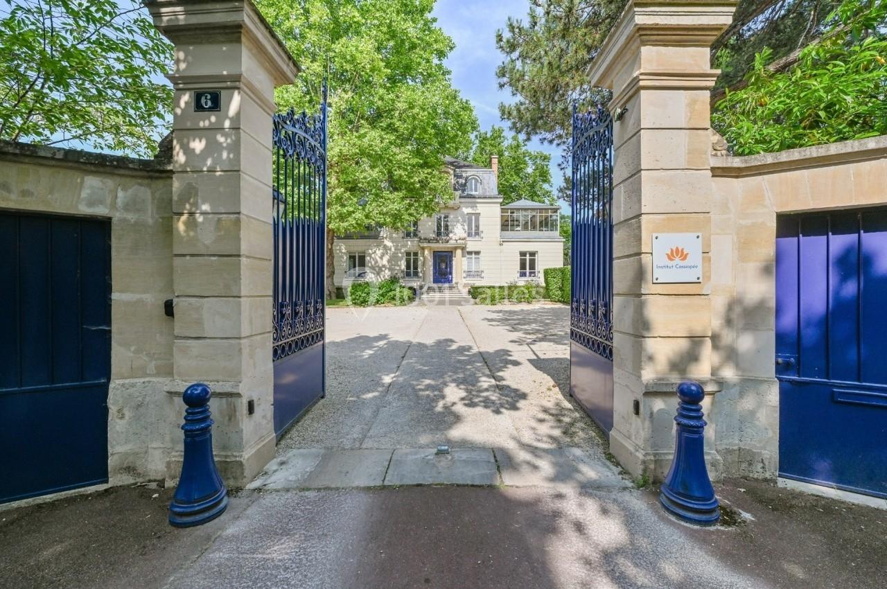 Entrée d'une propriété avec portail bleu ouvert, allée pavée et bâtiment en pierre entouré de verdure en arrière-plan.