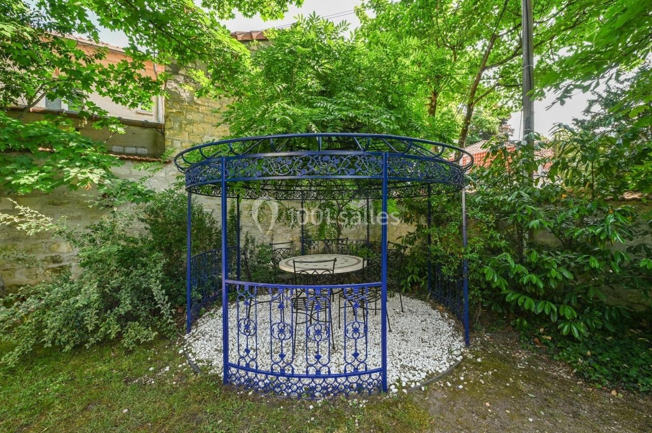 Tonnelle en fer forgé bleu entourée de végétation, abritant une table et des chaises sur un sol de gravier blanc.