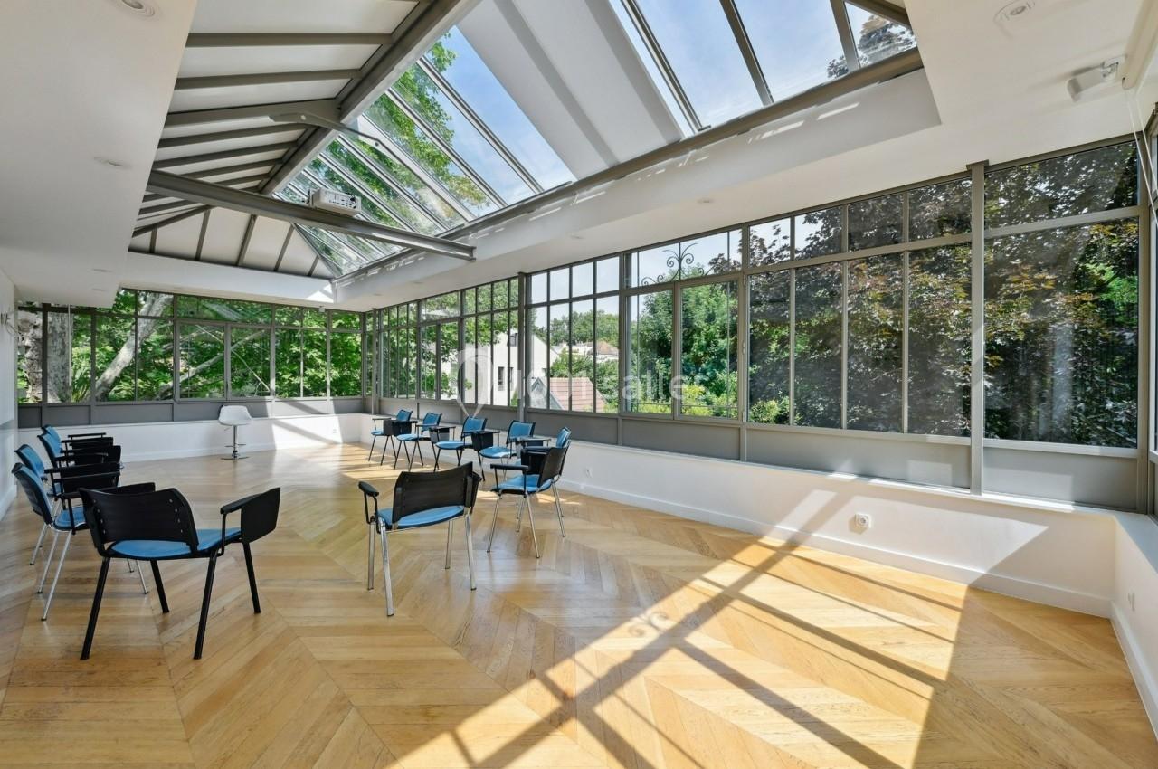 Salle lumineuse avec parquet en bois, baies vitrées et plafond en verrière, équipée de chaises disposées en cercle.