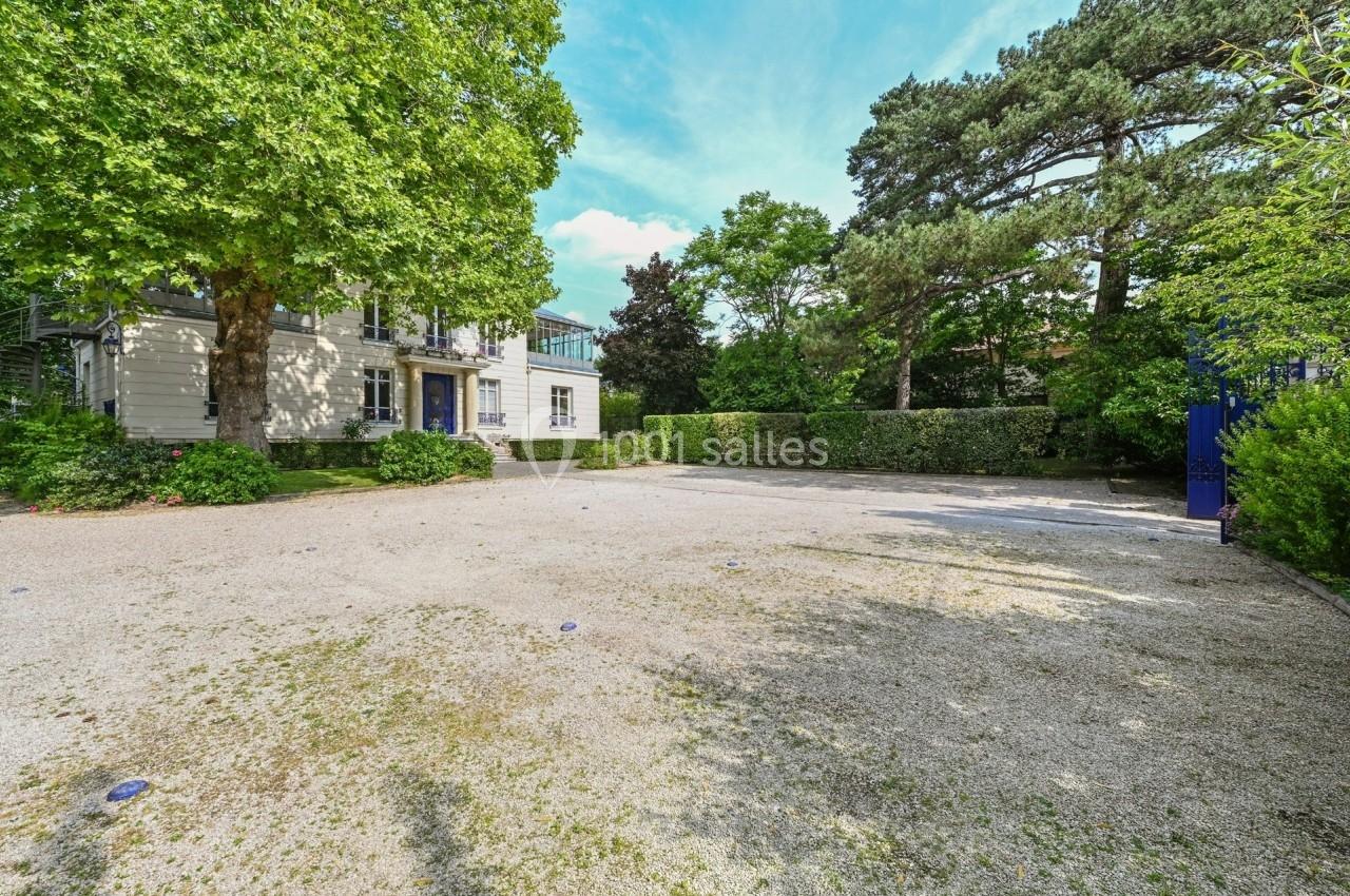 Cour gravillonnée entourée de verdure avec un bâtiment blanc à deux étages partiellement visible à gauche.