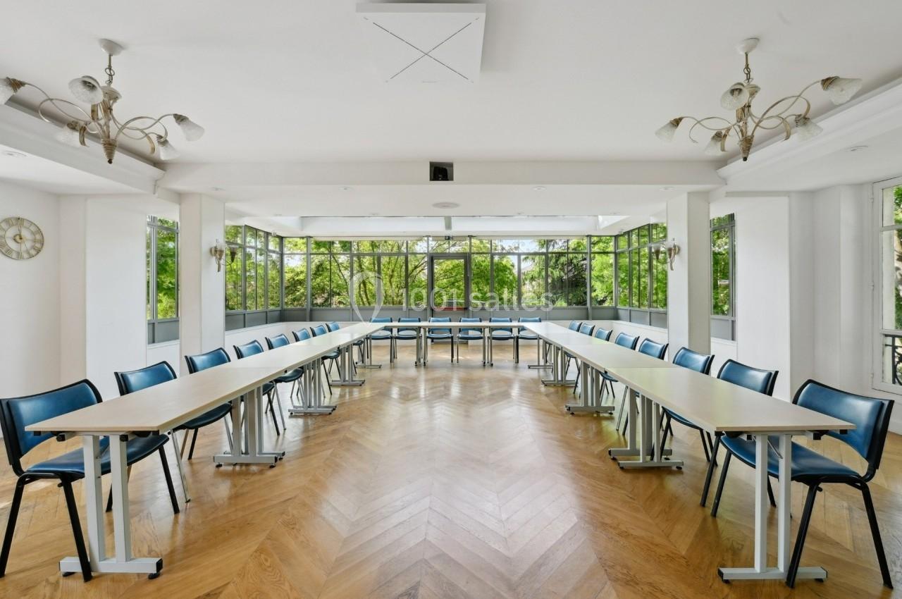 Salle lumineuse avec de grandes fenêtres, parquet en bois et tables disposées en U entourées de chaises.