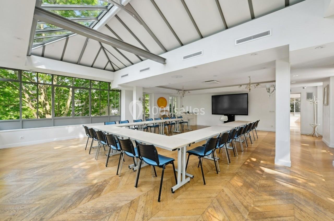 Salle lumineuse avec parquet en bois, grande table en U entourée de chaises, écran noir et verrière donnant sur des arbres.