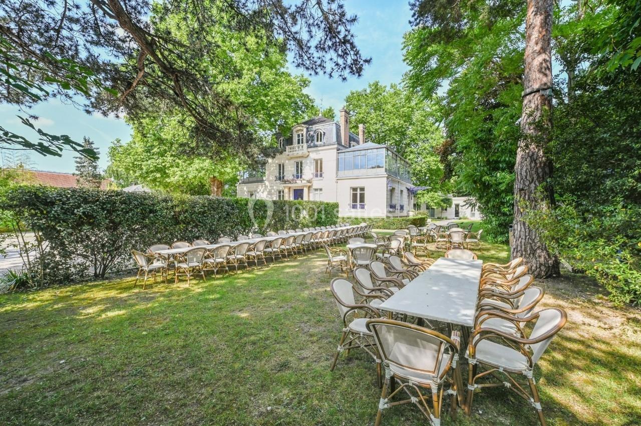Tables et chaises disposées en extérieur dans un jardin verdoyant, avec une grande maison en arrière-plan.