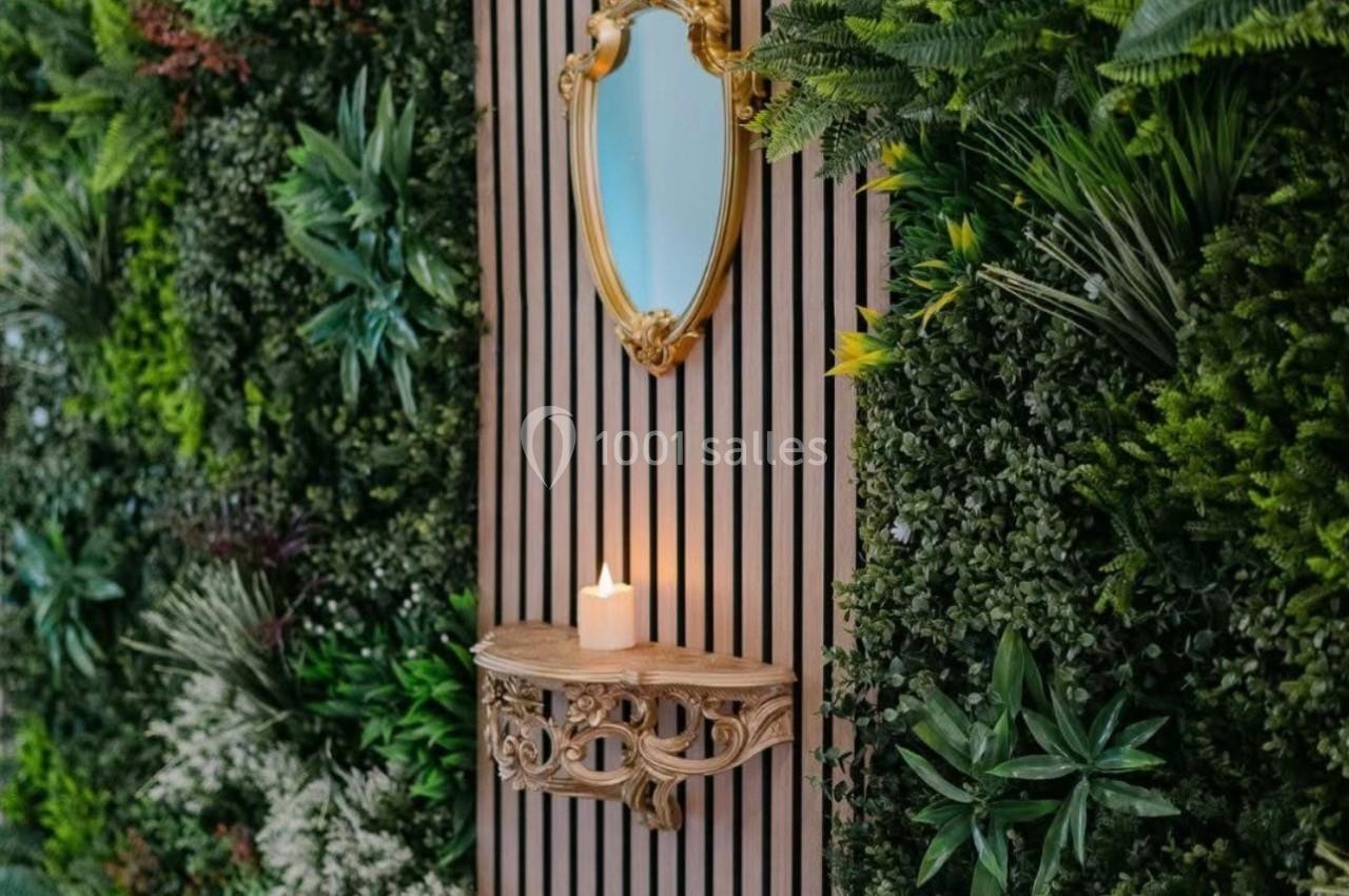 Mur végétal avec miroir doré ovale et bougie allumée posée sur une petite étagère sculptée en bois.