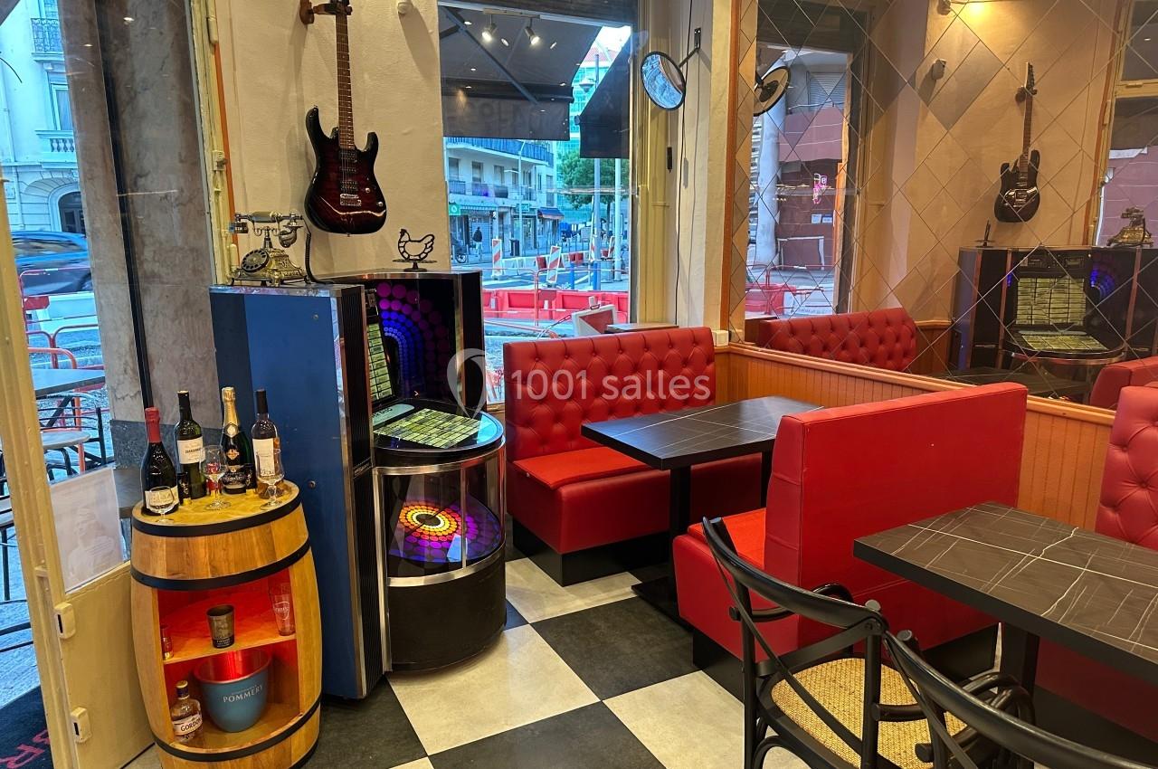 Intérieur d'un café avec banquettes rouges, tables noires, jukebox, guitare accrochée et vue sur la rue par la vitrine.