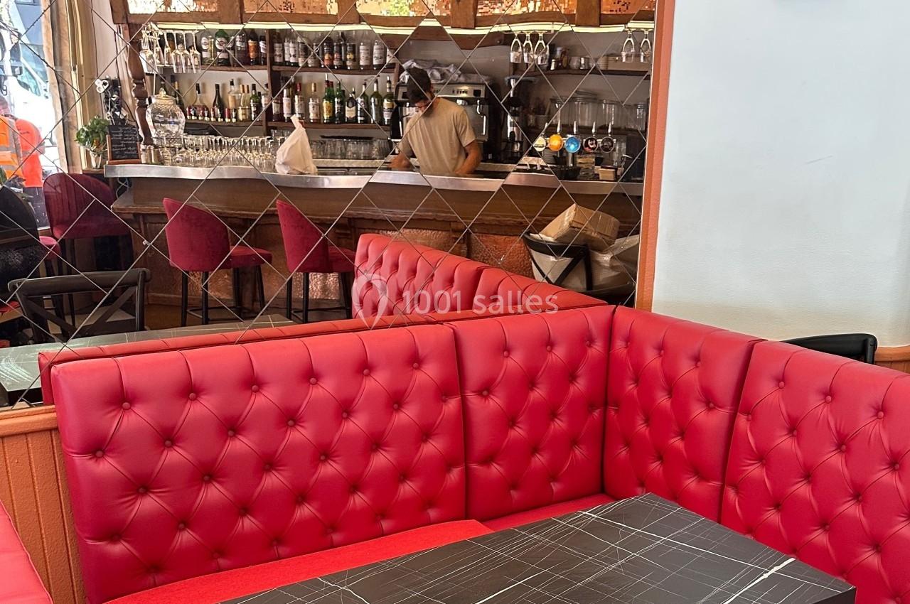 Banquette rouge capitonnée dans un café avec un bar en arrière-plan et une personne préparant des boissons.