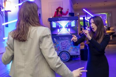 Deux personnes discutent gestuellement lors d'une soirée avec une DJ en arrière-plan et des lumières colorées.