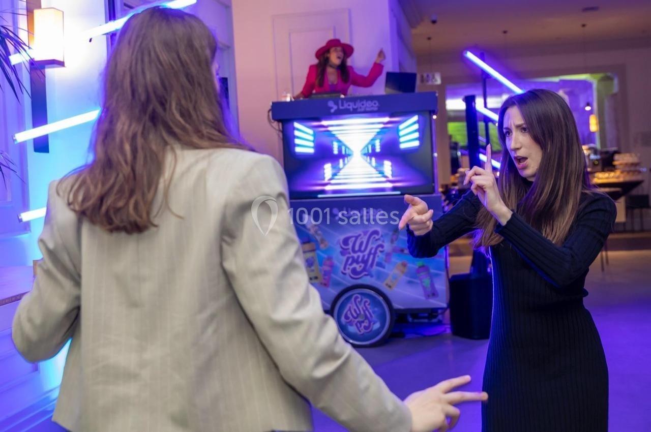 Deux personnes discutent gestuellement lors d'une soirée avec une DJ en arrière-plan et des lumières colorées.