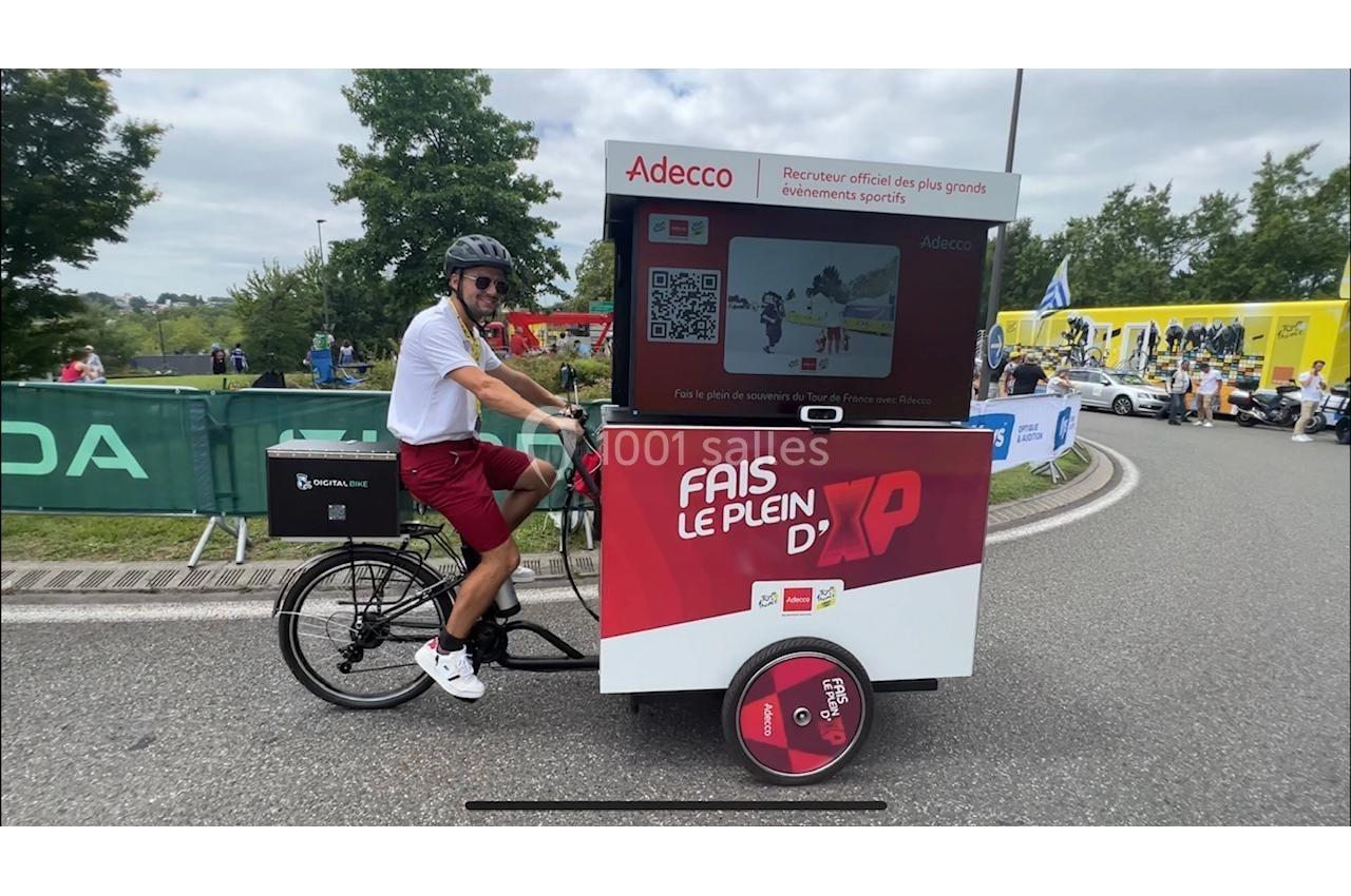 Un homme conduit un vélo cargo publicitaire Adecco sur une route bordée d'arbres et de spectateurs.
