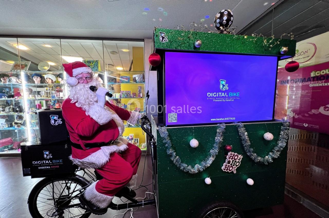 Un Père Noël assis sur un vélo décoré, tenant un micro devant un écran lumineux dans un espace intérieur.