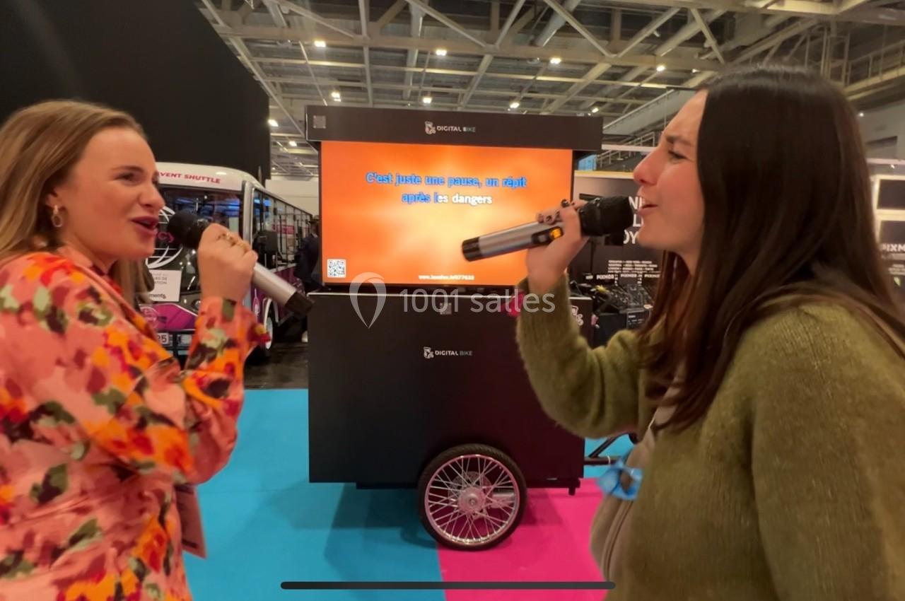 Deux femmes chantant au karaoké dans un espace d'exposition, tenant chacune un micro devant un écran.