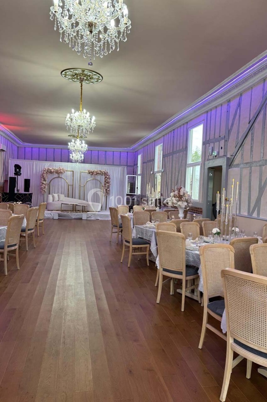 Salle de réception élégante avec tables dressées, chaises en osier, lustres et arche décorée de fleurs.