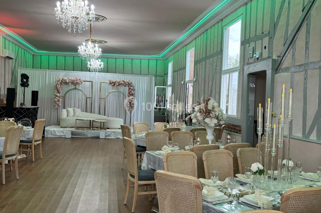 Salle de réception élégante avec tables dressées, chandeliers, décor floral et canapé sur une estrade.