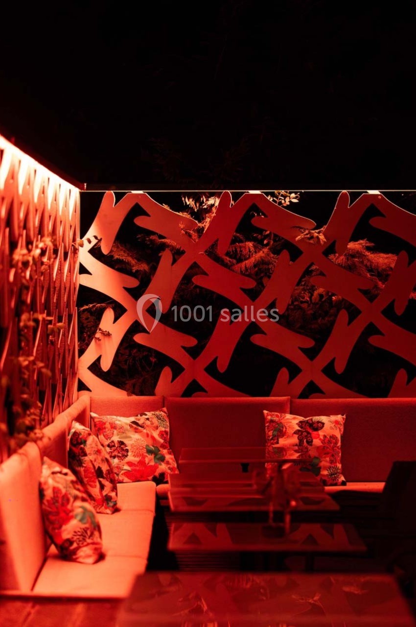 Espace extérieur éclairé en rouge, avec banquettes, coussins à motifs floraux et cloison décorative ajourée.