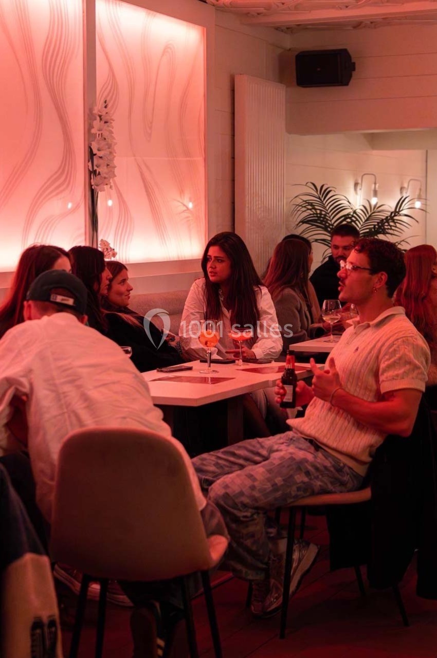 Des personnes discutent et partagent des boissons autour de tables dans un espace intérieur à l'éclairage tamisé rouge.