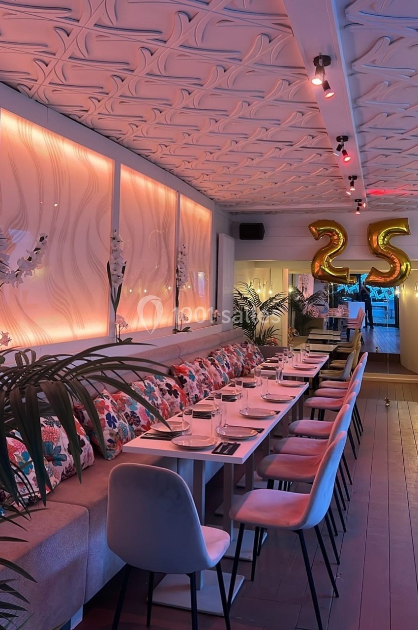 Salle de restaurant élégante avec une longue table dressée, décorée de coussins colorés et ballons dorés en forme de ’25’.