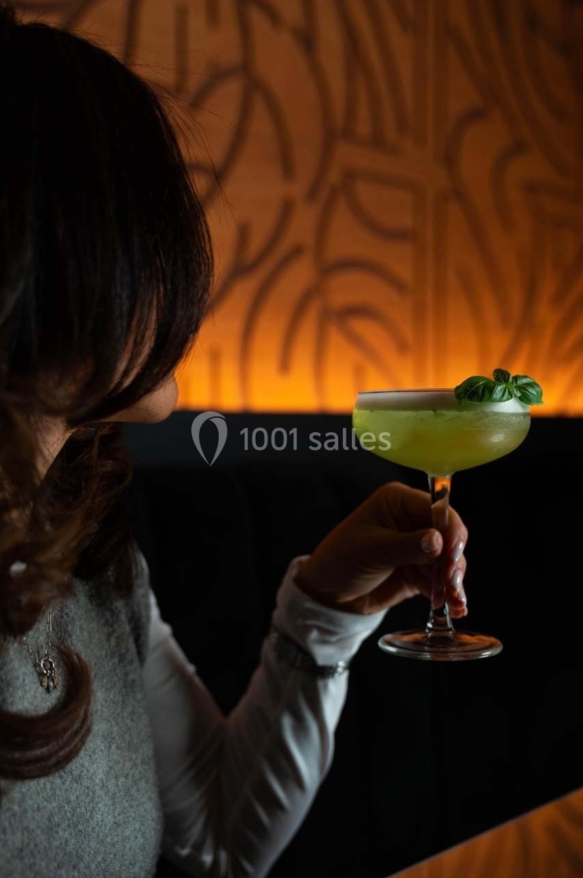 Une personne tenant un cocktail vert garni de feuilles de basilic, dans un cadre tamisé avec un mur texturé en arrière-plan.