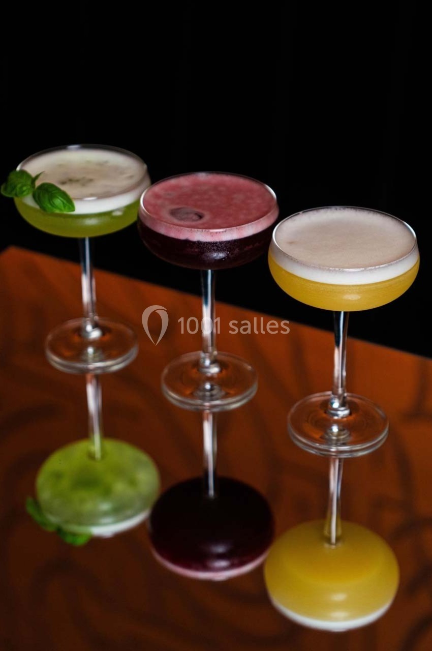 Trois cocktails colorés dans des verres à pied sur une surface réfléchissante, avec un fond sombre.