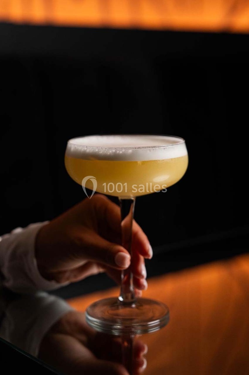 Un verre à cocktail rempli d'une boisson mousseuse jaune, tenu par une main sur une table sombre.