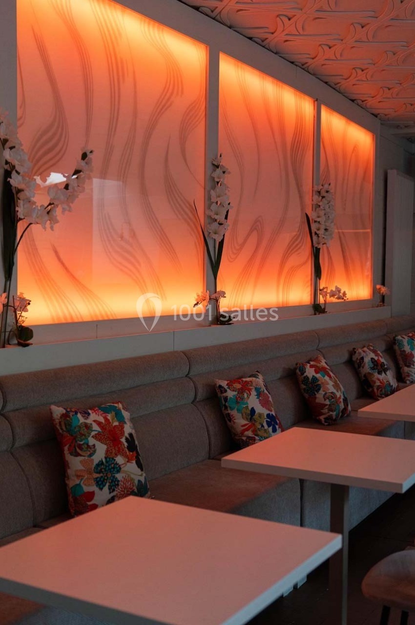 Banquettes grises avec coussins colorés, tables blanches et éclairage mural orange à motifs ondulés.