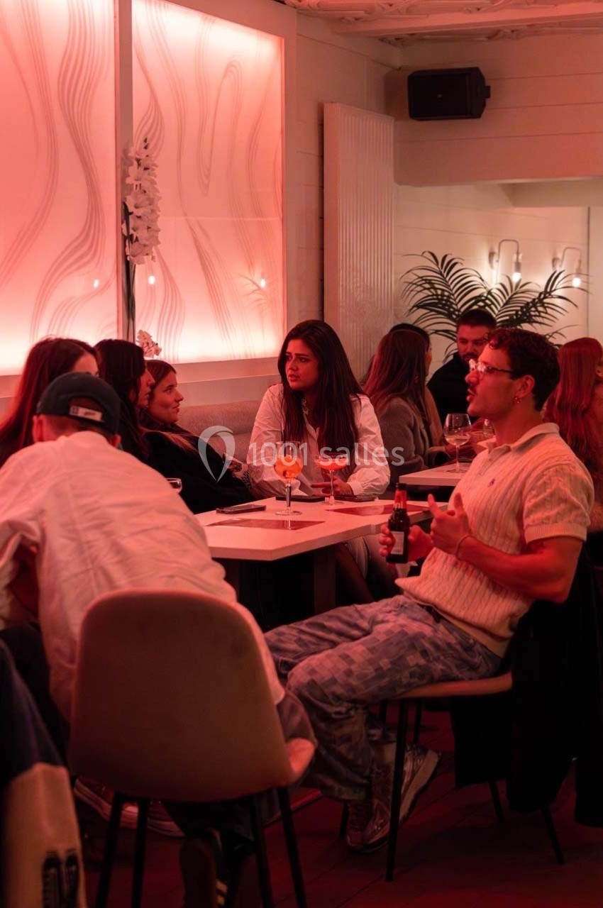 Des personnes assises autour de tables dans un restaurant à l'éclairage tamisé rose, discutant et partageant des boissons.