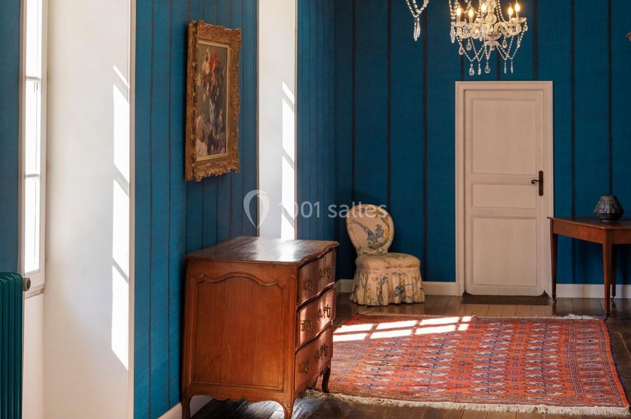 Intérieur lumineux avec murs bleus, commode en bois, fauteuil, tableau encadré, tapis rouge et lustre en cristal.