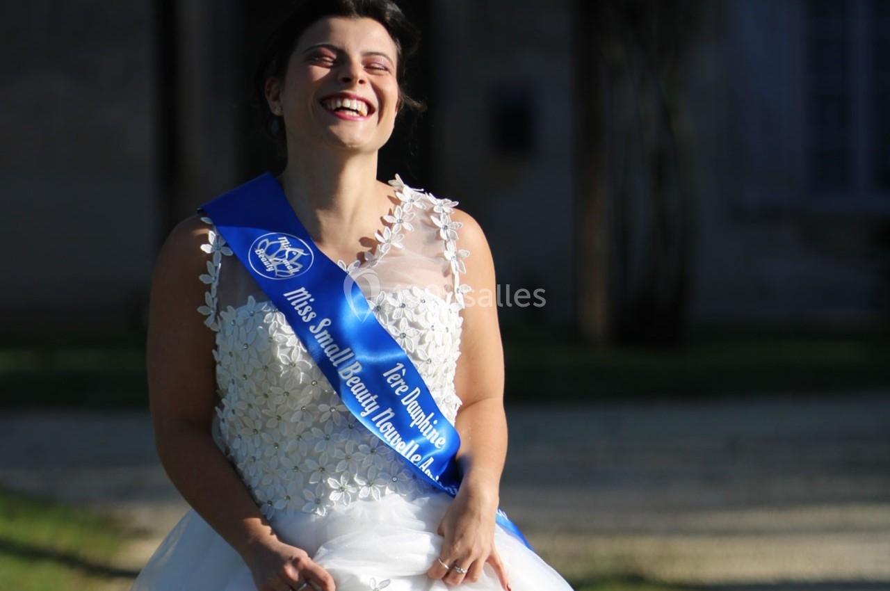 Femme souriante en robe blanche et écharpe bleue, debout à l'extérieur sous une lumière naturelle.