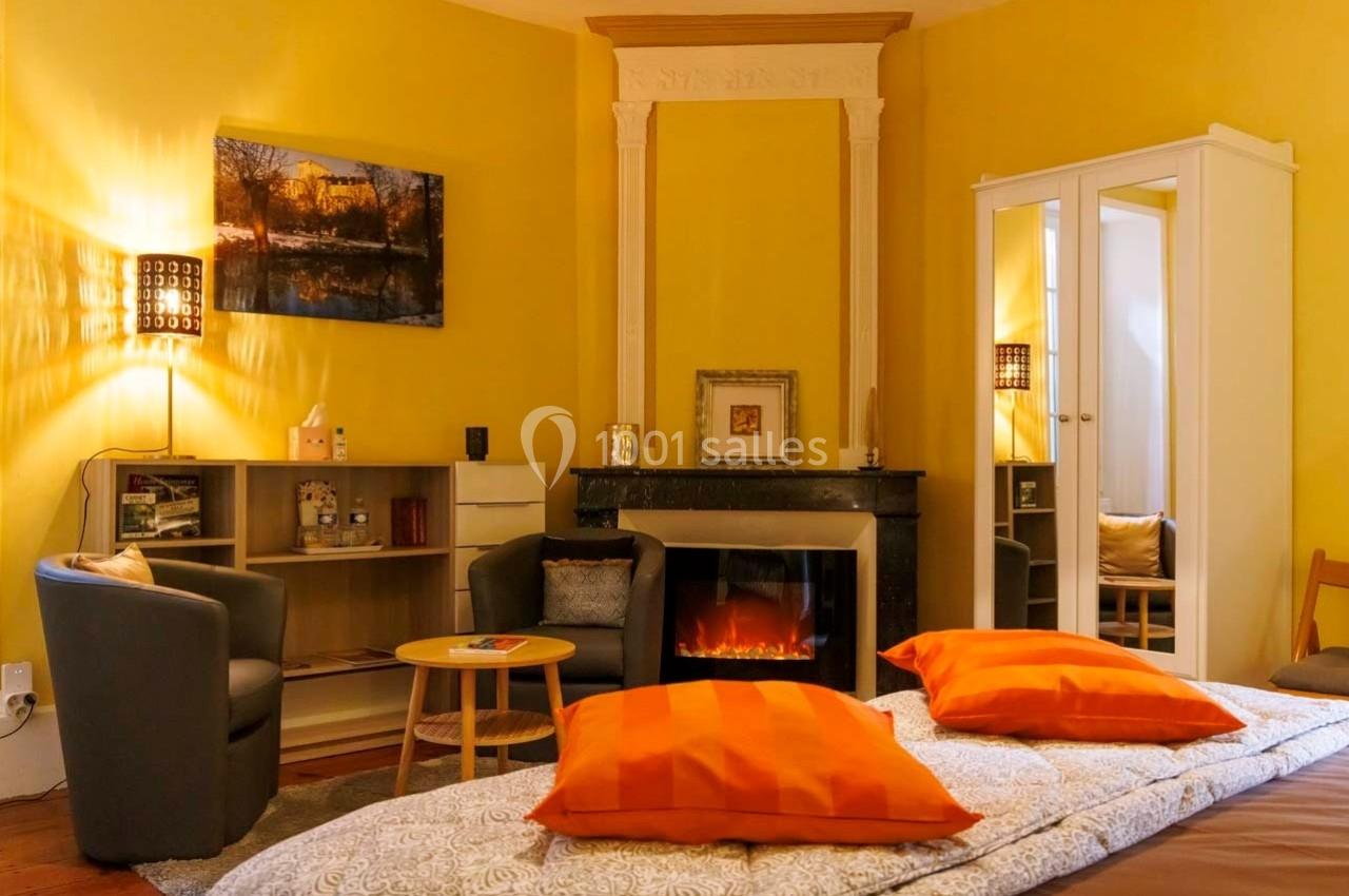 Chambre lumineuse avec murs jaunes, cheminée décorative, fauteuils gris, lit avec coussins orange et armoire blanche.