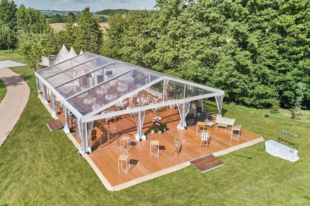 Grande tente transparente installée sur une plateforme en bois dans un espace extérieur verdoyant.