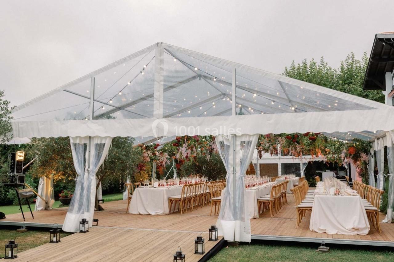 Grande tente transparente installée sur une terrasse en bois, décorée de guirlandes lumineuses et de fleurs, avec tables…