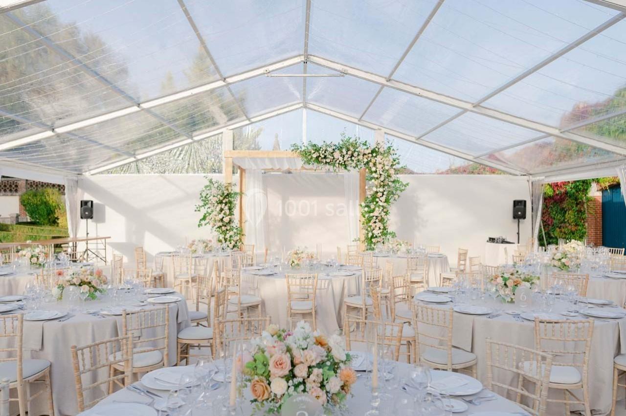 Salle de réception sous une tente transparente, décorée de fleurs et tables rondes dressées avec élégance.