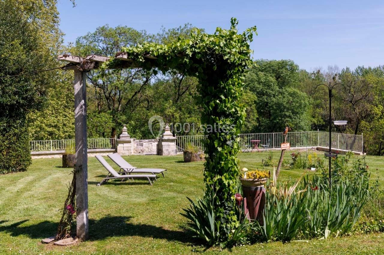 Pergola couverte de lierre dans un jardin verdoyant avec des chaises longues et une vue sur des statues en pierre.