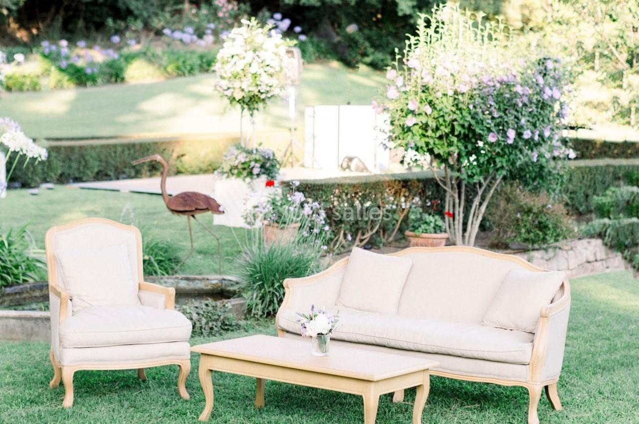 Mobilier de jardin beige avec canapé, fauteuil et table basse, installé sur une pelouse entourée de plantes et fleurs.