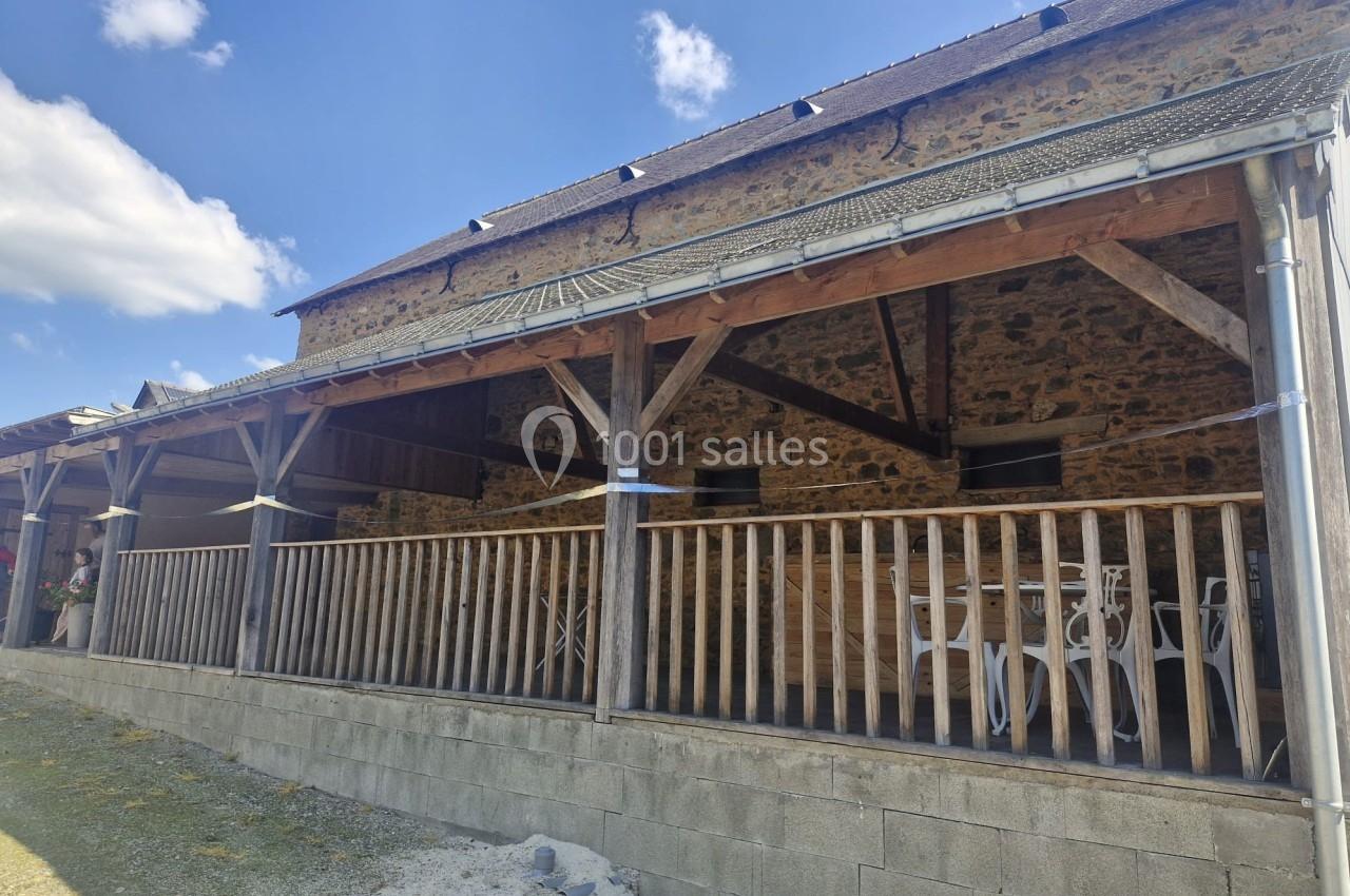 Terrasse couverte en bois d'une bâtisse en pierre, avec des chaises blanches visibles sous l'abri.
