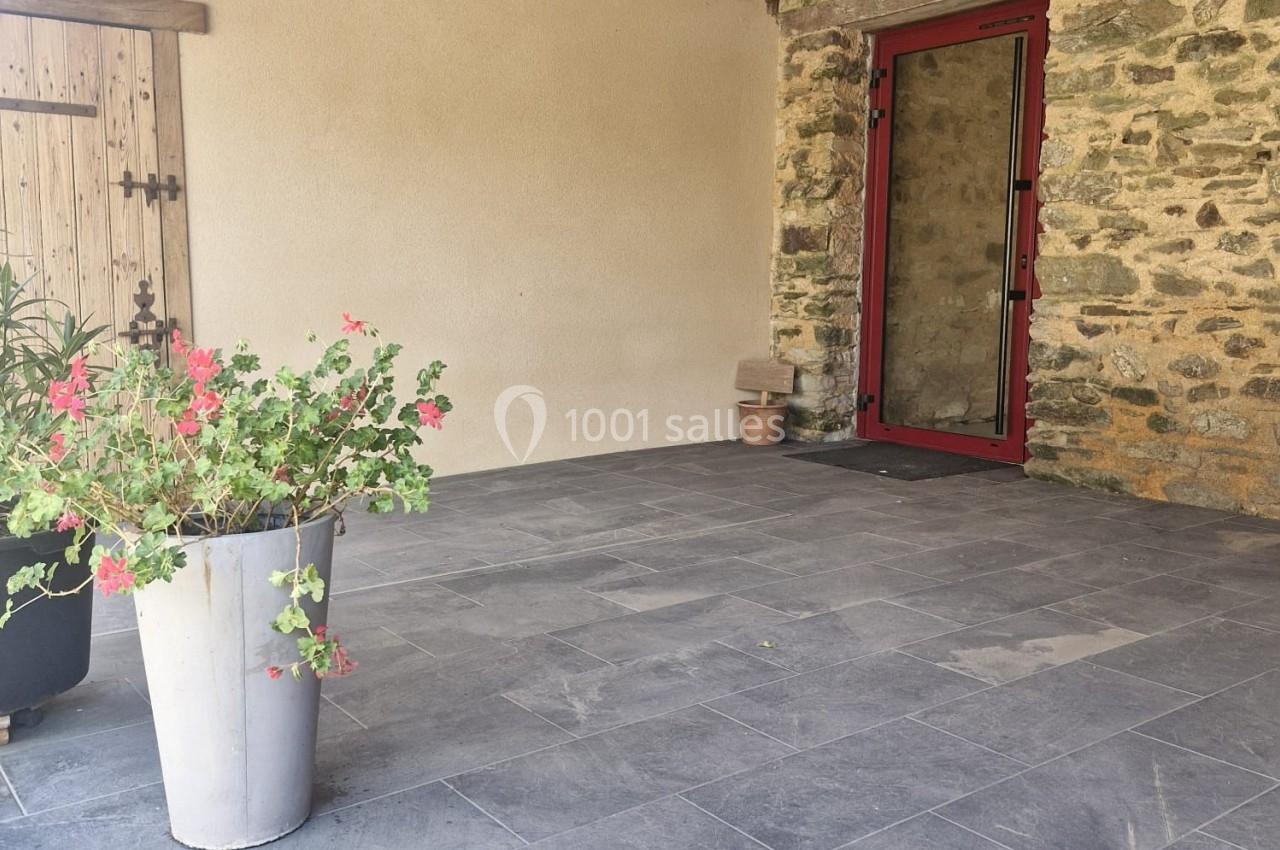 Terrasse couverte avec sol en carrelage gris, mur en pierre, porte vitrée rouge et plantes en pots.