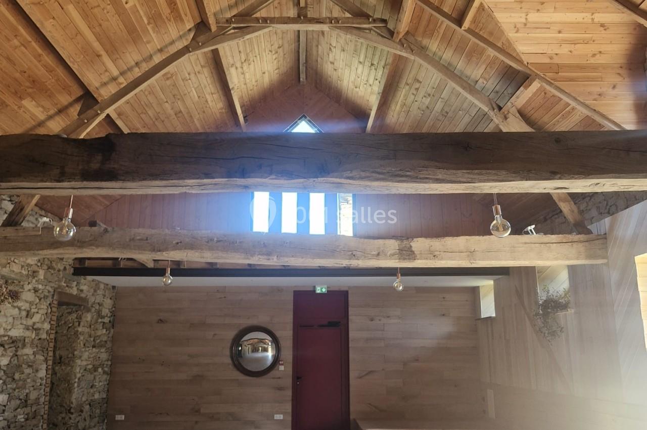 Salle intérieure lumineuse avec plafond en bois, poutres apparentes, murs en pierre et porte rouge au fond.