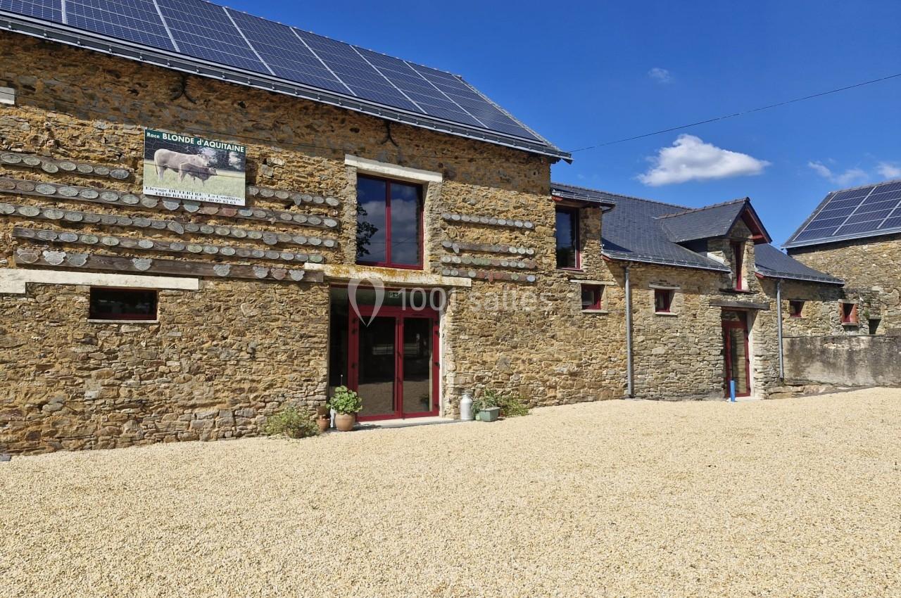 Façade en pierre d'un bâtiment rural avec panneaux solaires sur le toit, entouré d'une cour en gravier.