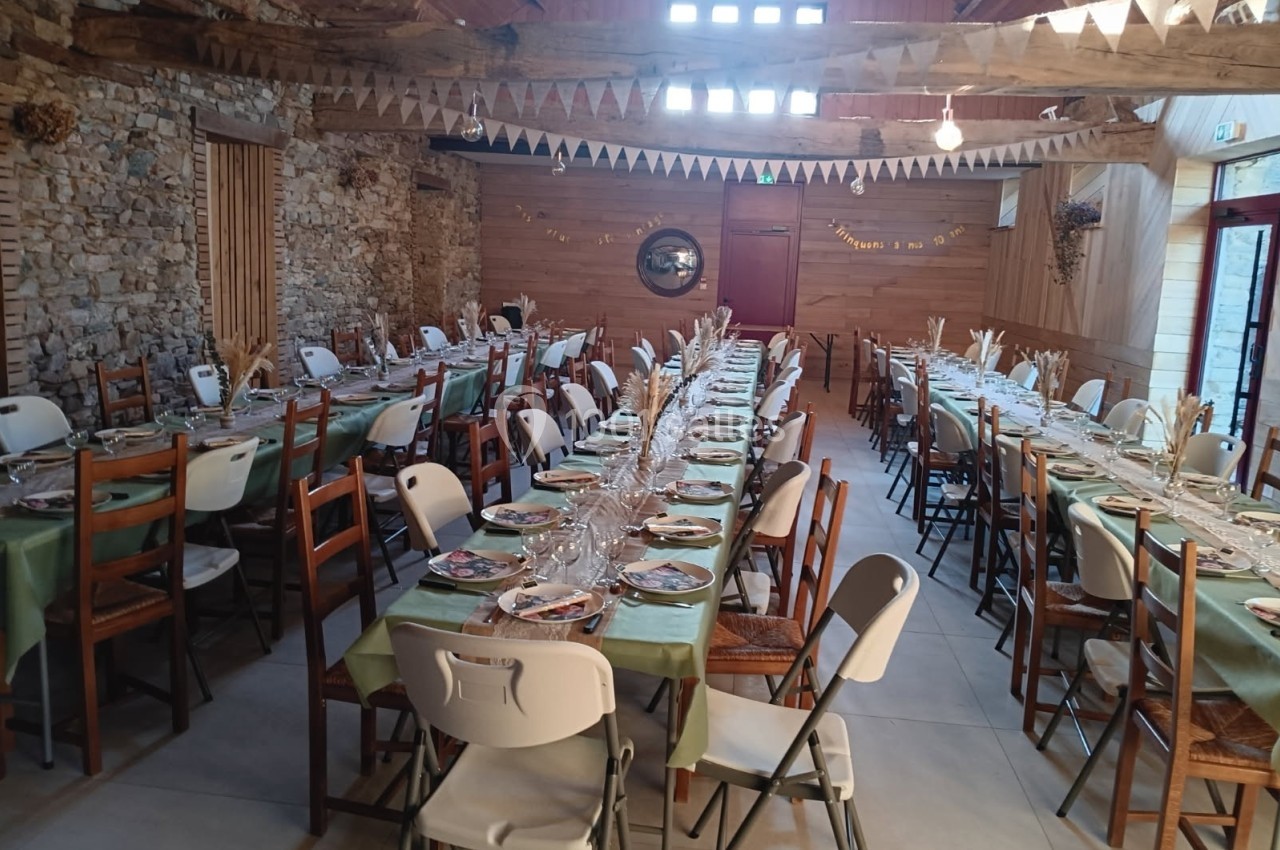 Salle de réception décorée avec des tables longues dressées, nappes vertes et guirlandes suspendues sous un plafond en bois.