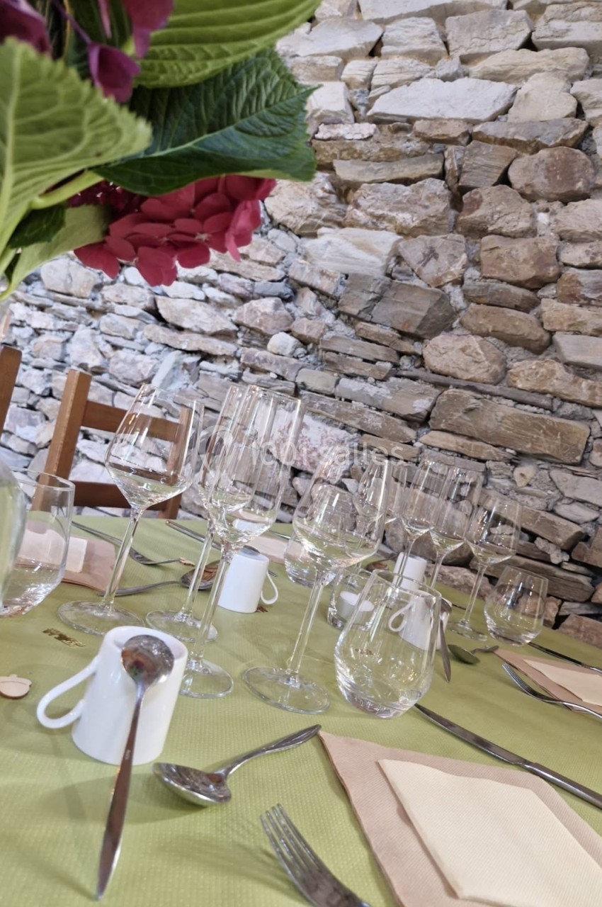 Table dressée avec nappes vertes, verres à vin, couverts, serviettes et un vase de fleurs devant un mur en pierre.