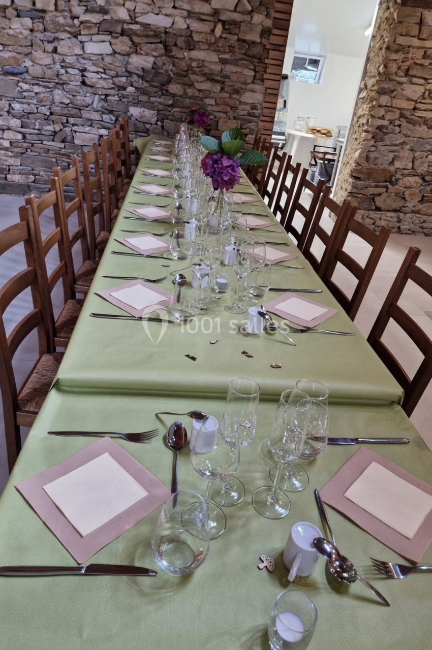 Table dressée avec une nappe verte, couverts, verres et serviettes, dans une salle aux murs en pierre apparente.