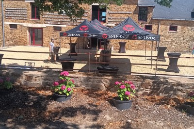 Cour extérieure ensoleillée avec des tentes noires, des fleurs en pot et une maison en pierre en arrière-plan.