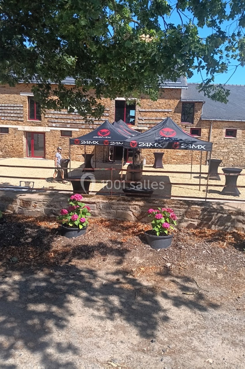 Cour extérieure ensoleillée avec des tentes noires, des fleurs en pot et une maison en pierre en arrière-plan.