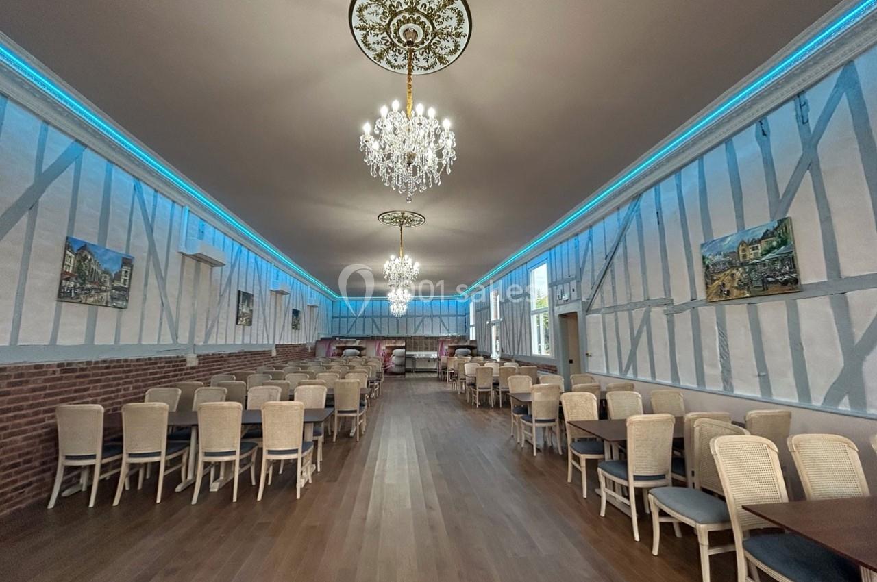 Grande salle lumineuse avec parquet en bois, murs à colombages, lustres élégants et chaises alignées autour de tables.