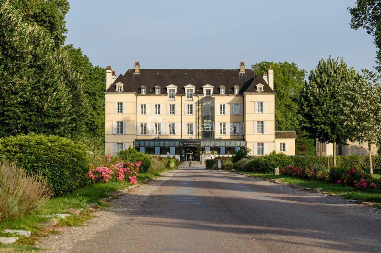 Façade d'un grand bâtiment de style classique entouré de jardins fleuris et d'une allée bordée d'arbres.