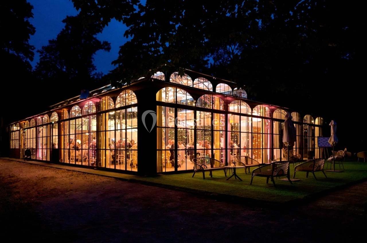 Grande serre illuminée de nuit, avec des invités visibles à l'intérieur et des chaises disposées sur la pelouse devant.