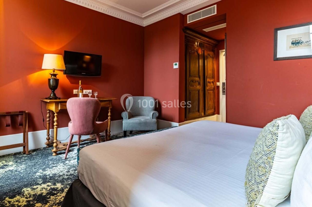 Chambre d'hôtel avec lit double, bureau en bois, fauteuils, mur rouge et éclairage chaleureux.
