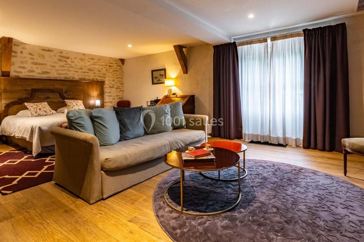 Chambre lumineuse avec lit double, canapé, table basse et rideaux sombres devant une grande fenêtre.