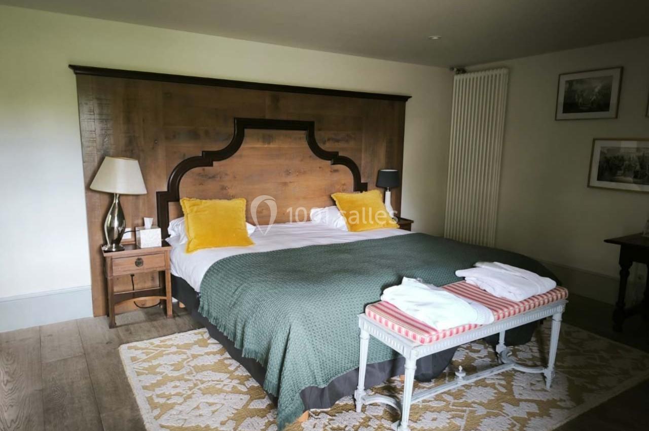 Lit double avec tête de lit en bois, coussins jaunes, banc avec serviettes pliées et éclairage doux dans une chambre.