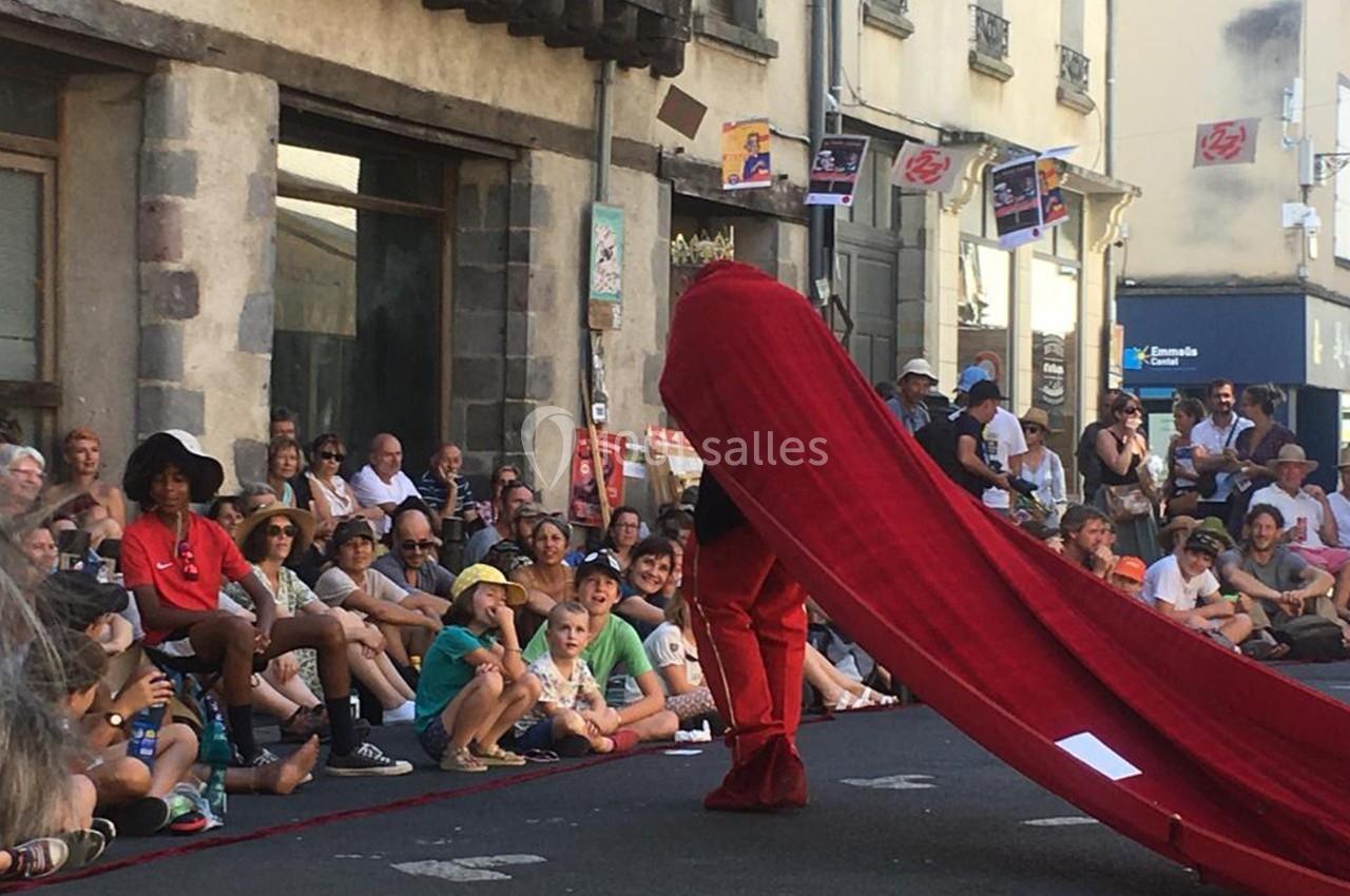 Un artiste en costume rouge avec une longue cape se produit devant un public assis dans une rue animée.